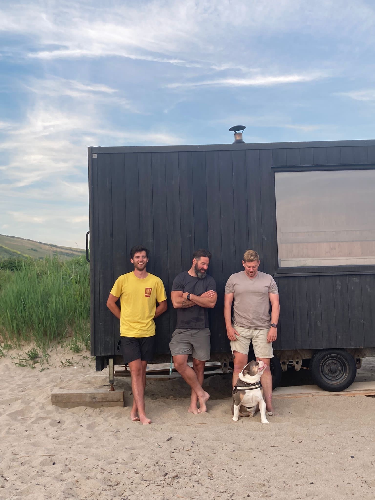 Three men stand barefoot on the sand in front of a wooden trailer