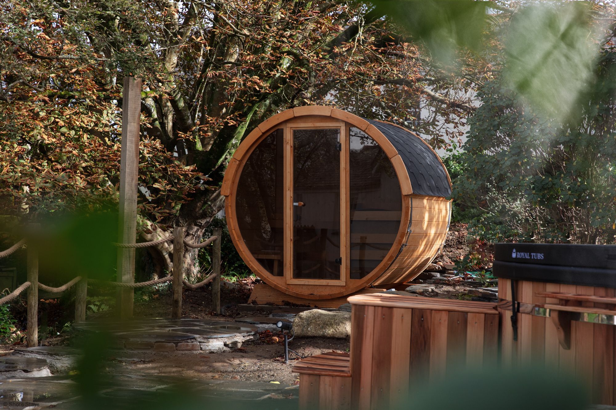 A wooden barrel-shaped sauna with a glass door amongst autumn trees.