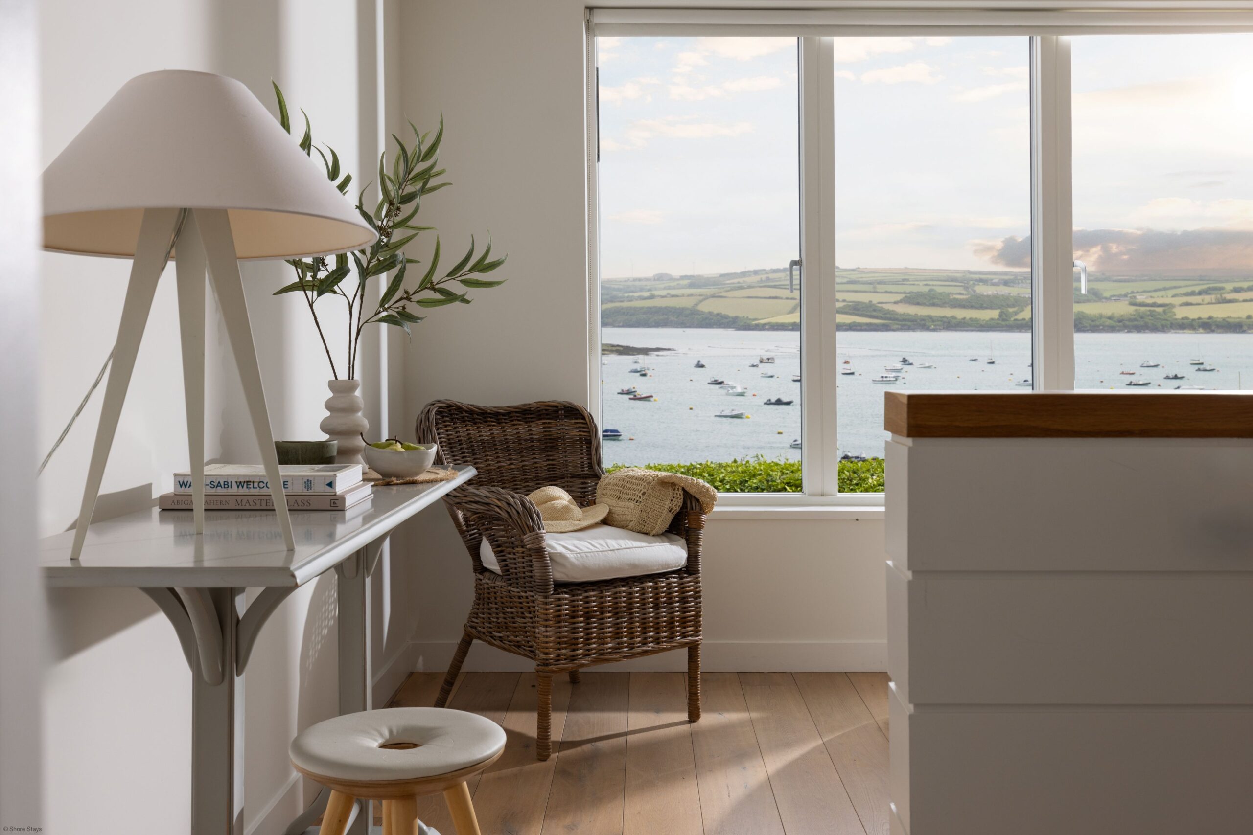 A room with a wicker chair and table lamp, facing a large window with a view of the sea.