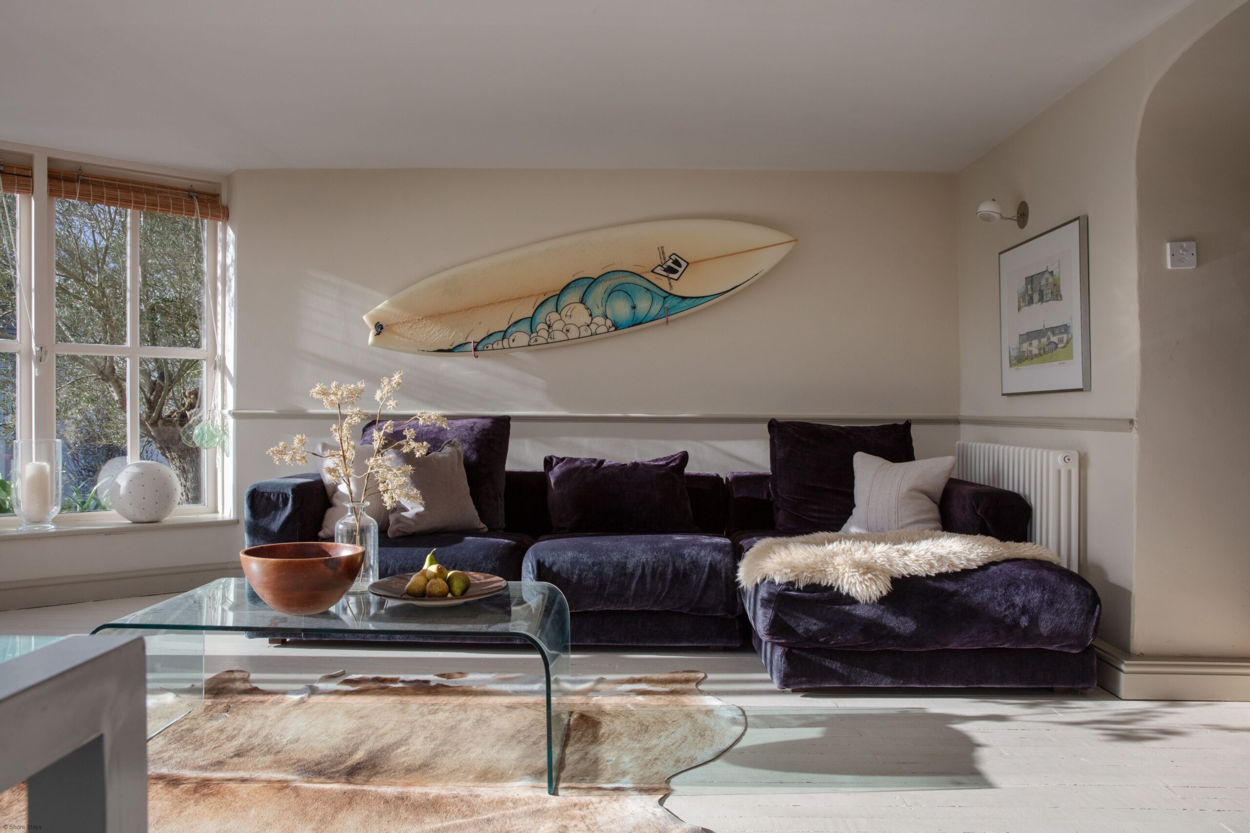 A living room with a dark velvet sectional sofa, surfboard wall art, and a glass coffee table holding a bowl and fruit.