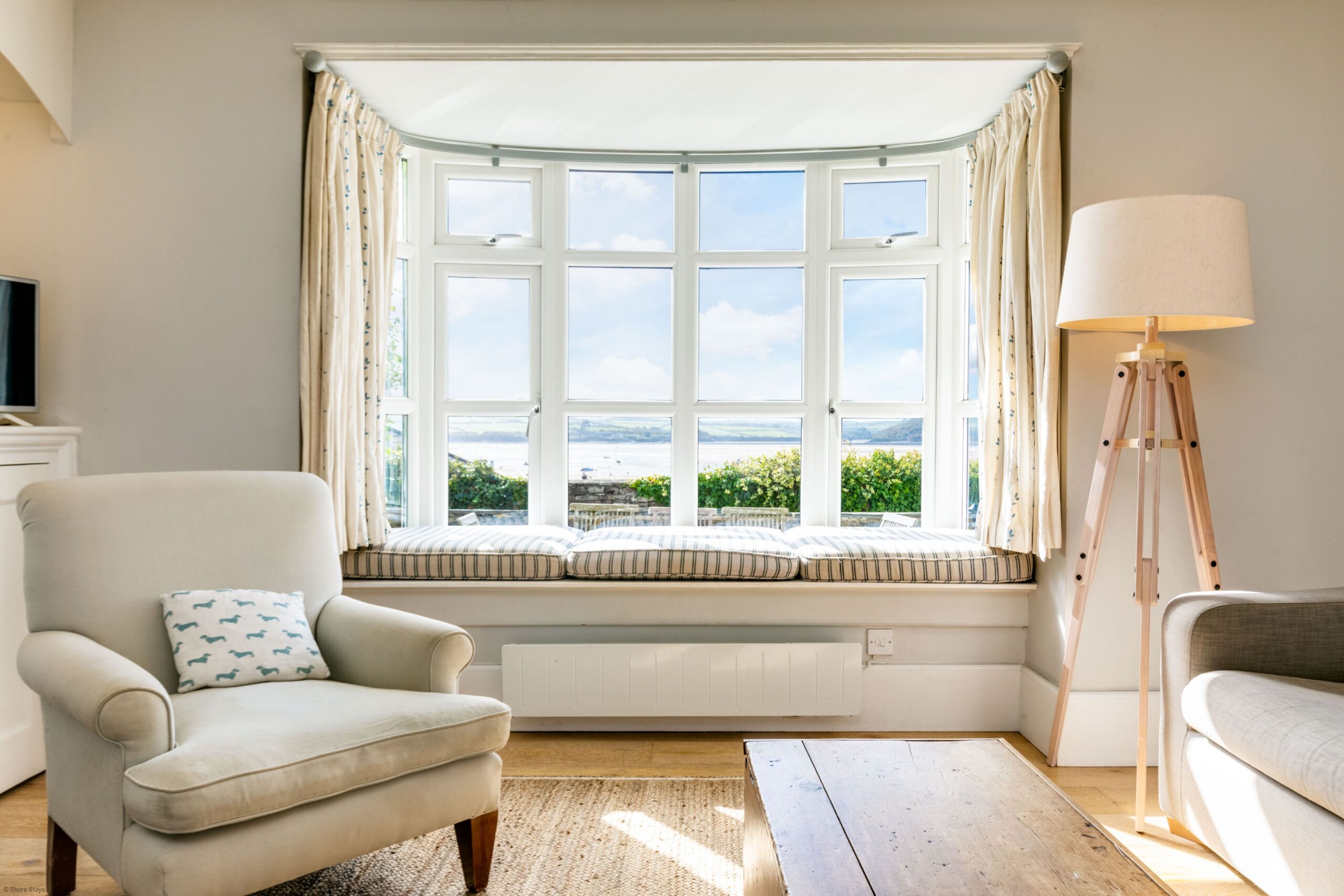 Bright living room with an armchair and cushion, wooden coffee table, tall floor lamp, and large bay window.