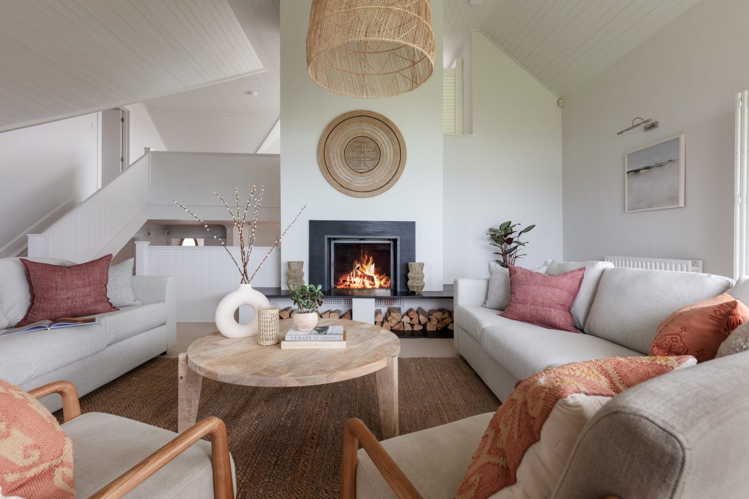 A living room with a lit fireplace, surrounded by light grey sofas with pink and orange cushions