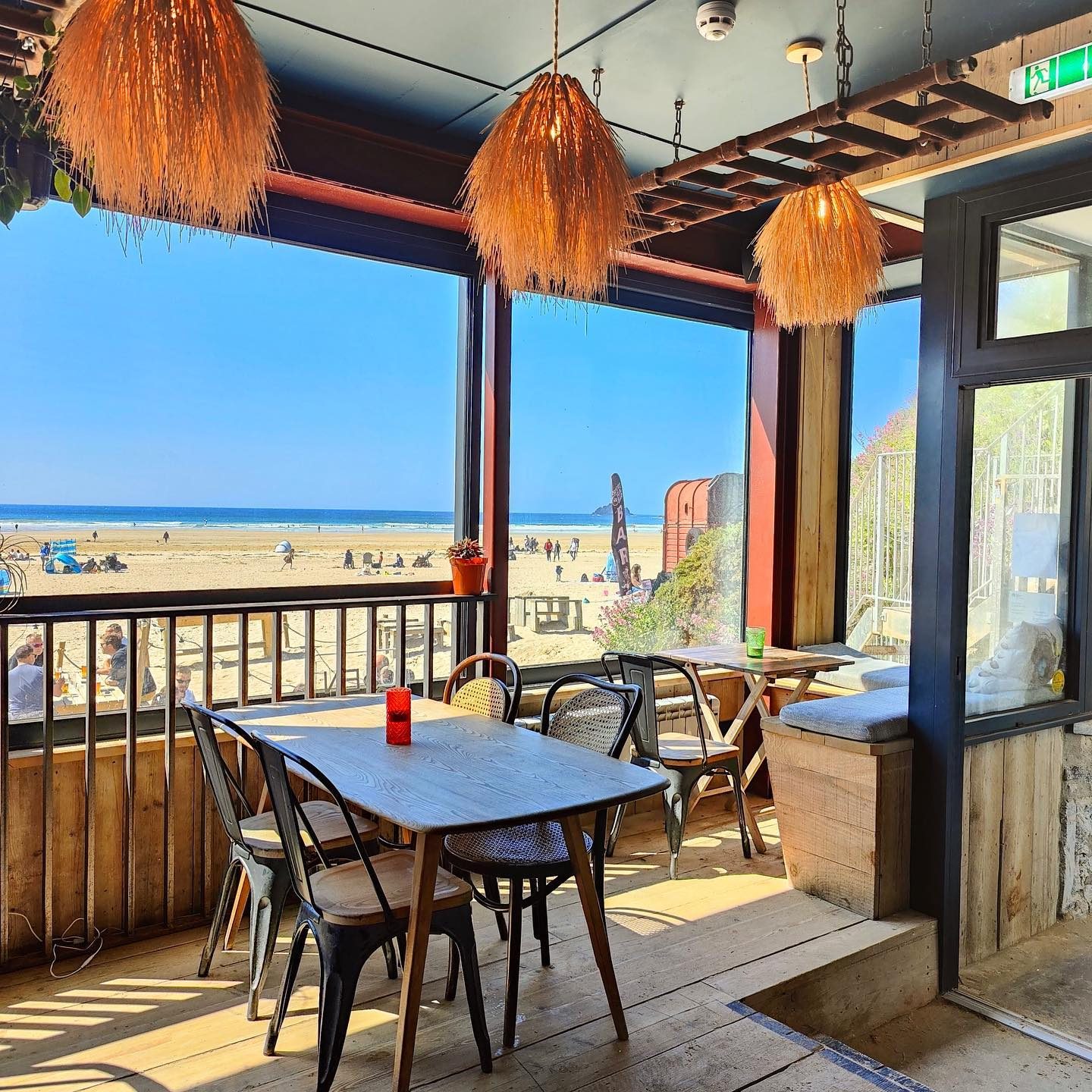 A beachside cafe with wooden tables, wicker chairs, and woven pendant lights