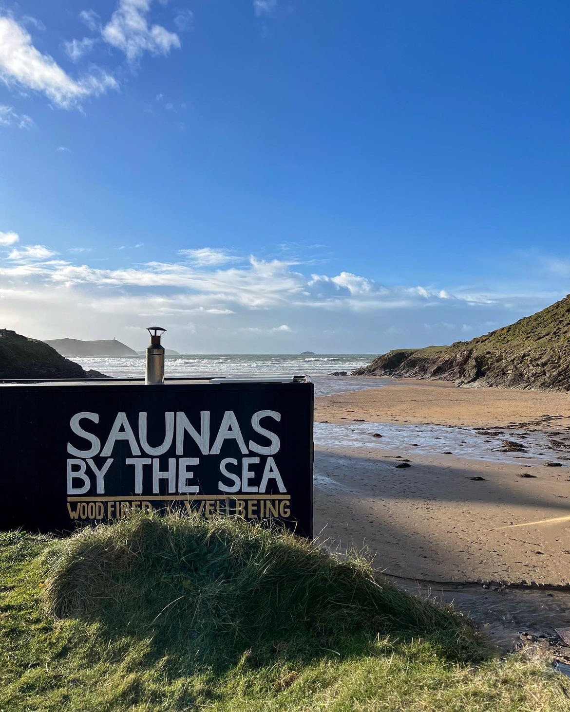 A beach on a sunny say with a wooden trailer reading "Saunas by the Sea"
