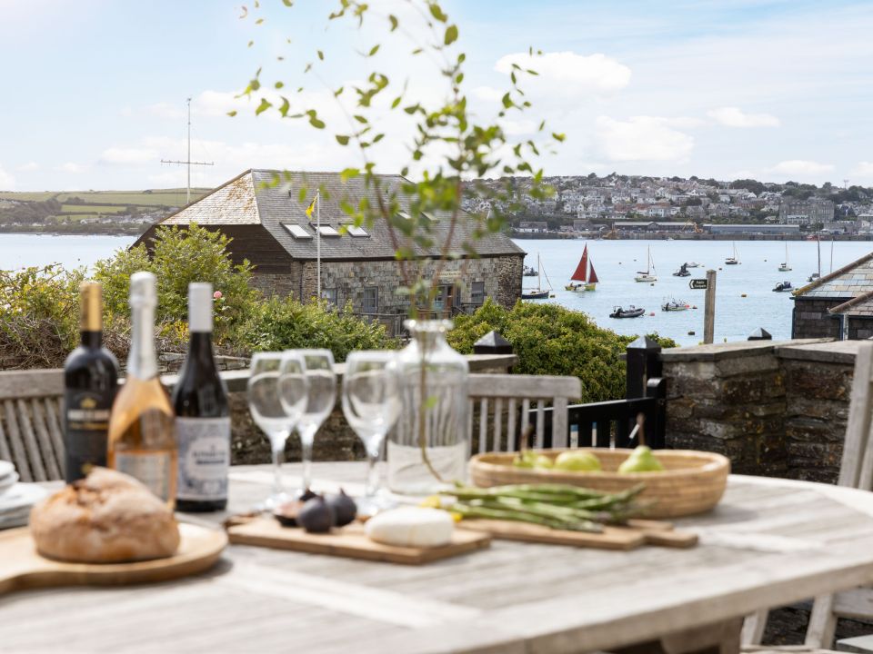 Outdoor table set with wine, glasses, bread, figs, and pears, overlooking a harbour.