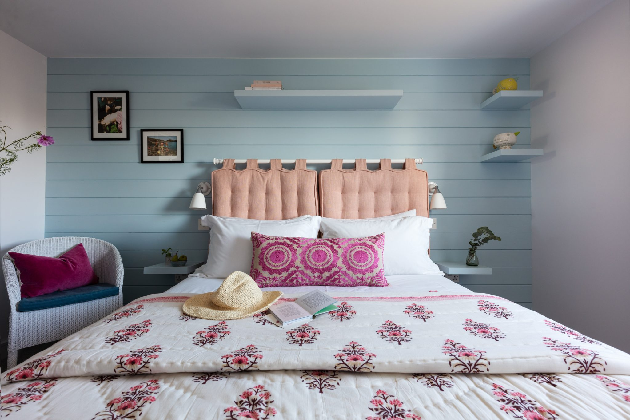 A bedroom with pastel blue walls, a pink tufted headboard, floral bedspread, and decorative shelves.