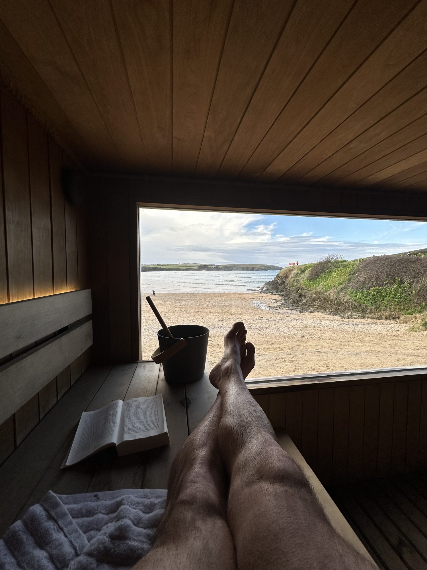 Person relaxing in a wooden sauna room, legs stretched out, with an open book and bucket nearby.