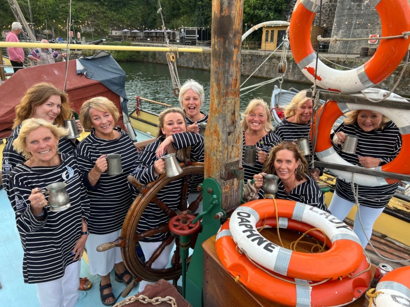 Nine women in striped tops on a boat, smiling and holding metal mugs