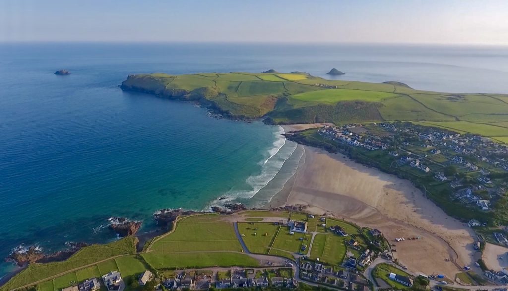 Aerial view of a coastal village with a sandy beach and blue ocean, bordered by green fields.