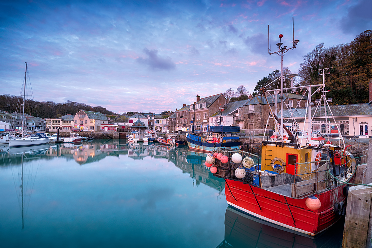 Padstow harbour at dawn with still water reflecting boats and traditional buildings.