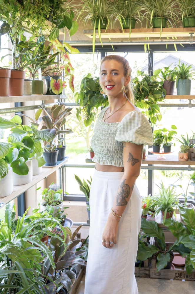 A woman smiling and standing amongst indoor plants on wooden shelves