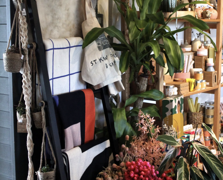 A shop corner with a black ladder displaying textiles, a tote bag, and potted plants