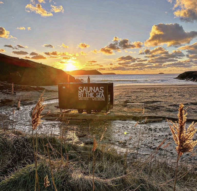 Sunset over a beach with a small trailer building labelled "Saunas by the Sea."