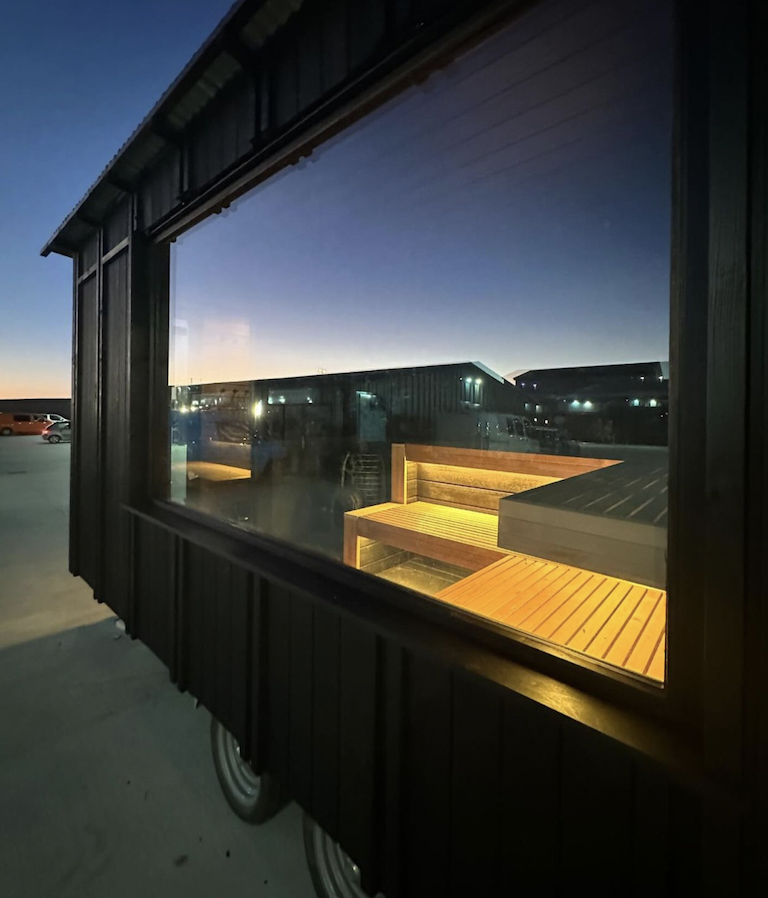 Reflection of a glowing yellow sauna interior in a large window at dusk.