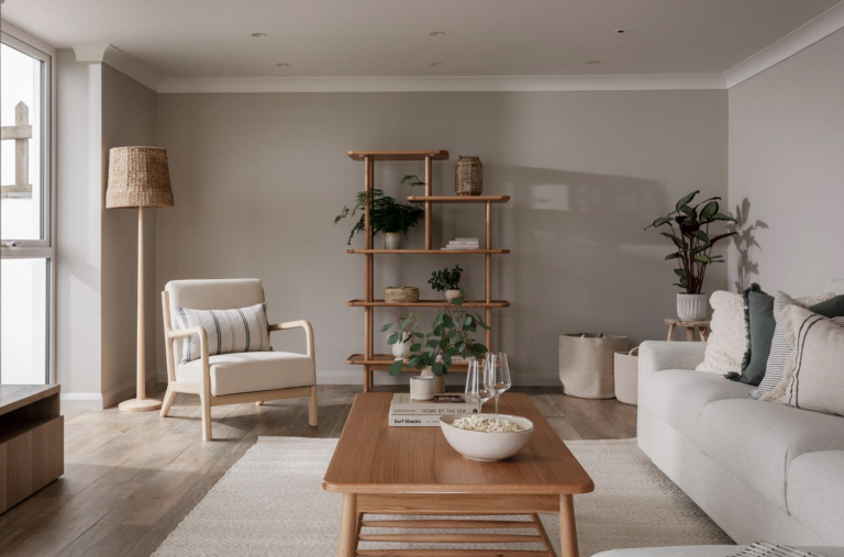 A living room with a light wooden coffee table, beige armchair, modern sofa, tall bookshelf, and potted plants.