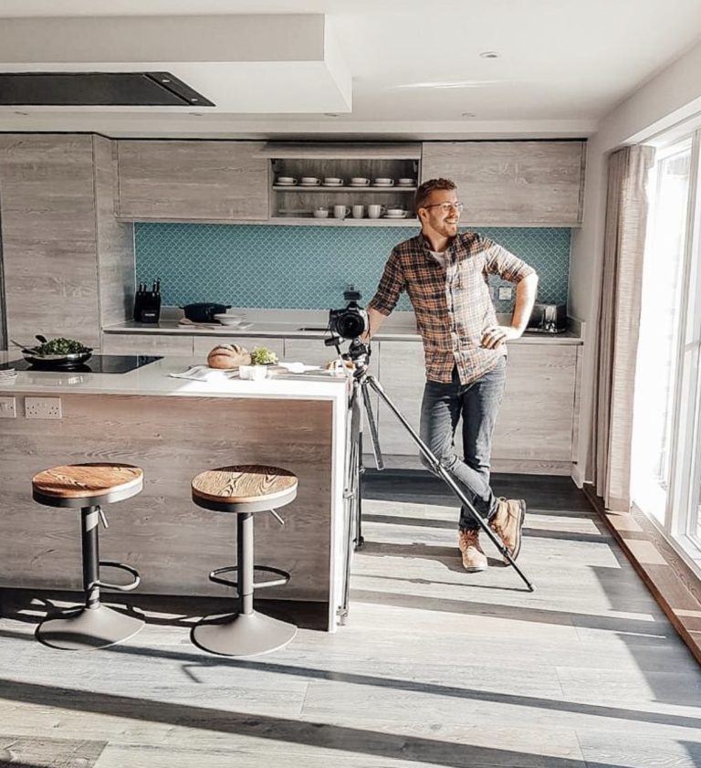 A man leaning on a camera tripod in a bright kitchen