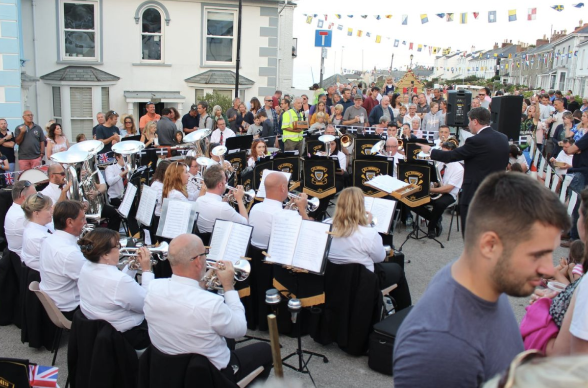 A brass band in white shirts and black vests performing outdoors, surrounded by a large audience.