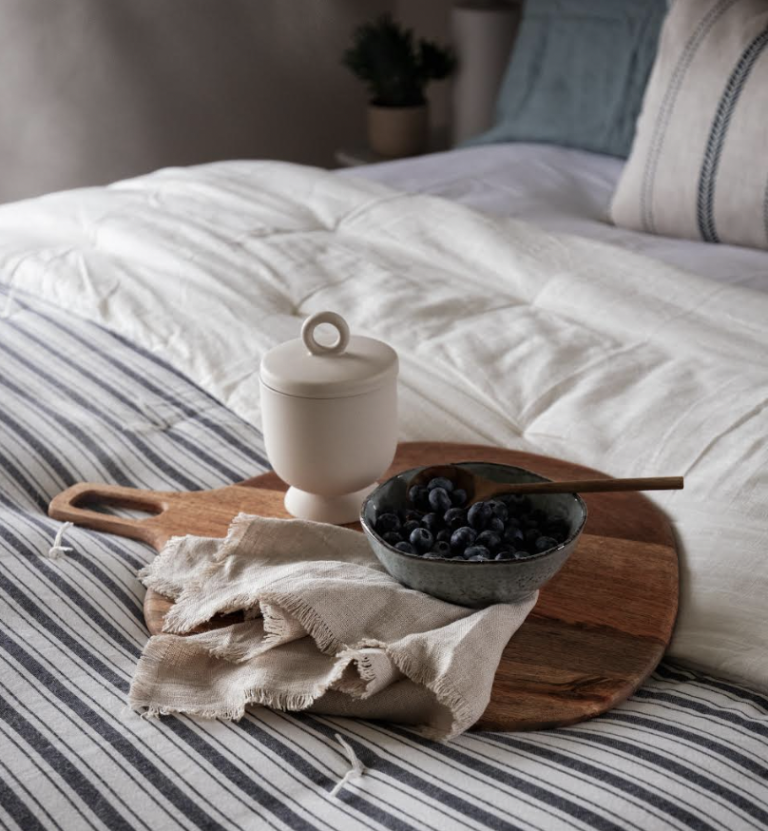 A wooden tray holding a bowl of blueberries, a white sugar pot, and a beige cloth on striped bedding.