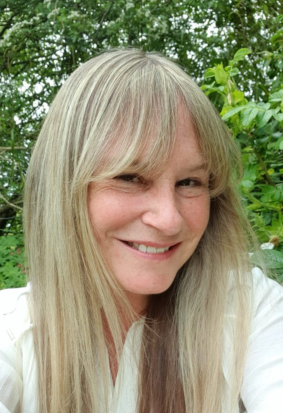 A smiling woman with blonde hair outdoors, surrounded by trees.