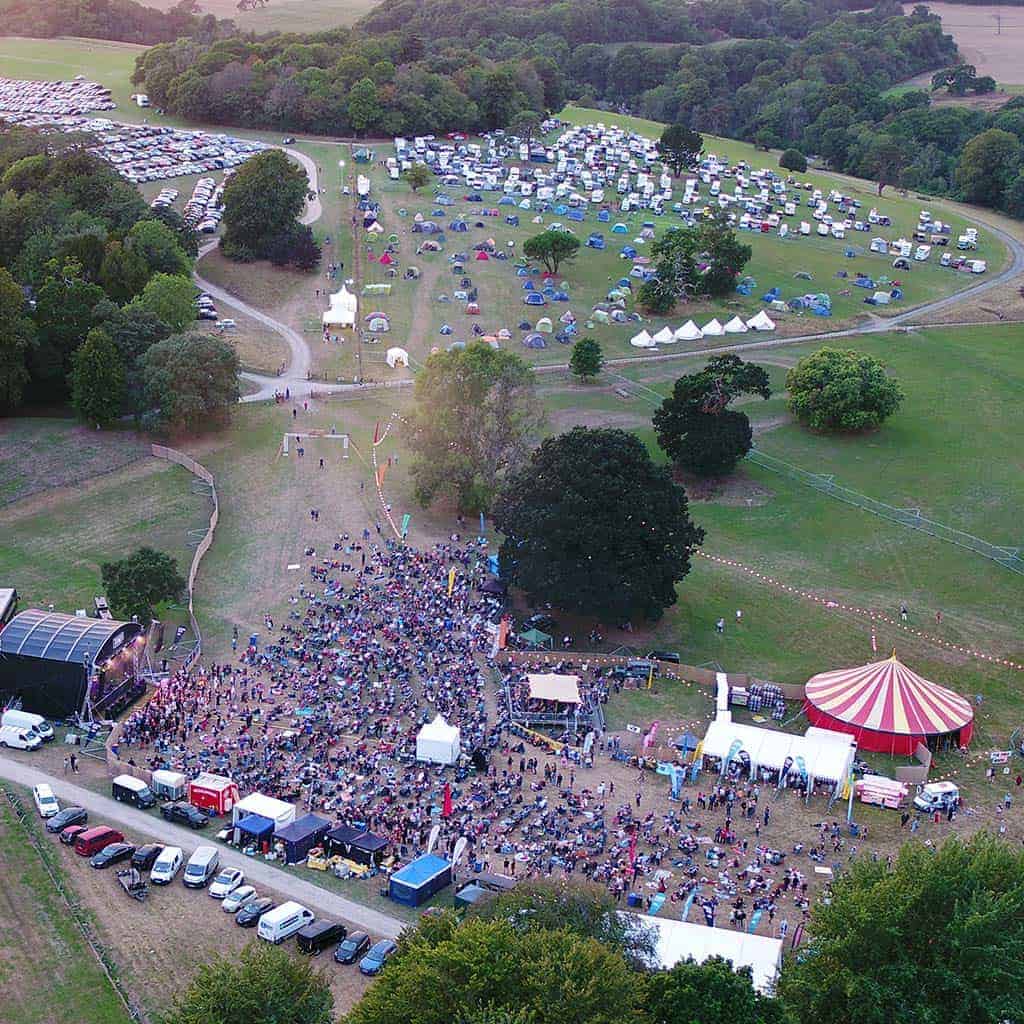 Aerial view of a vibrant outdoor festival with crowds near a stage, colourful tents, food trucks, and parked cars